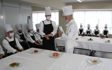 会場の会議室の机に白い布。そこにサラダが並んでいる。生徒たちが周りに座り、発表する学生は先生の目の前に立ち、プレゼンをしている様子。